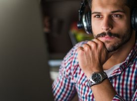 Man with dark hair and mustache wearing a gingham shirt with a watch, large over the ear headphones and looking at a grey computer monitor.