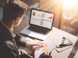 Young Finance Market Analyst In Eyeglasses Working At Sunny Office On Laptop While Sitting At Wooden Table.businessman Analyze Document In His Hands.graphs And Diagramm On Notebook Screen.blurred.