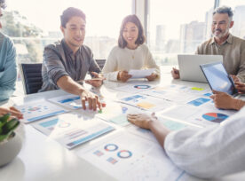 Multi Racial Diverse Group Of People Working With Paperwork On A Board Room Table At A Business Presentation Or Seminar. The Documents Have Financial Or Marketing Figures, Graphs And Charts On Them. There Are Laptops And Digital Tablets On The Table