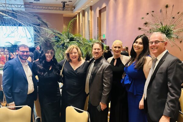 MCWT Peacock gala image of 8 people next to a table in a large event space. There are multicolored peacock feathers behind them.