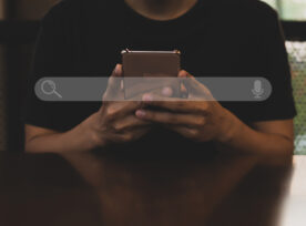 Young Asian Man Wearing Black T Shirt Sitting On Desk Hand Holding Smartphone To Searching For Information. Using Search Console With Your Website.