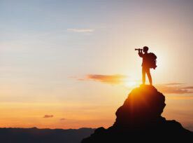 Silhouette Of Man Holding Binoculars On Mountain Peak Against Bright Sunlight Sky Background.