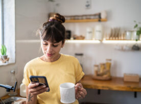 Young Woman Using The Mobile Phone While Drinking Coffee Or Tea At Home