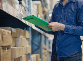 Asian Manager Man Doing Stocktaking Of Products Management In Cardboard Box On Shelves In Warehouse Using Clipboard File And Pen. Male Professional Assistant Checking Stock In Factory. Physical Inventory Count.