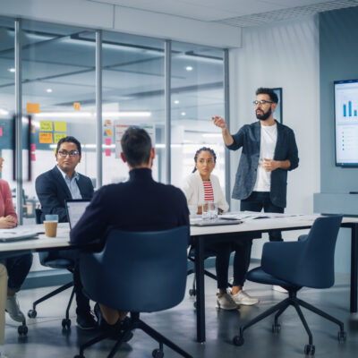 Diverse Modern Office: Businessman Leads Business Meeting With Managers, Talks, Uses Presentation Tv With Statistics, Infographics. Digital Entrepreneurs Work On E Commerce Project.