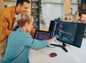 Smiling Male Programmer Explains A Coding Technique To A New Female Colleague While Sitting At A Desk In The Office