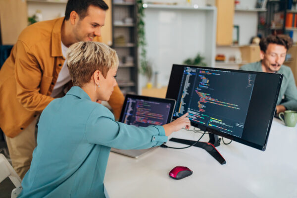 Smiling Male Programmer Explains A Coding Technique To A New Female Colleague While Sitting At A Desk In The Office