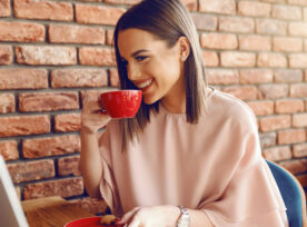 Portrait Of Attractive Young Caucasian Woman With Brown Hair And Toothy Smile Drinking Coffee While Sitting In Cafeteria And Looking At Laptop.