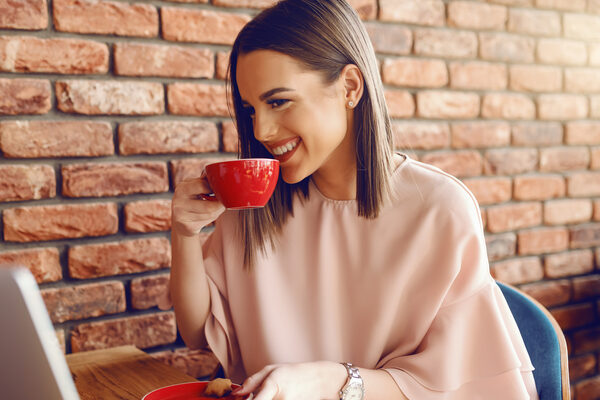 Portrait Of Attractive Young Caucasian Woman With Brown Hair And Toothy Smile Drinking Coffee While Sitting In Cafeteria And Looking At Laptop.