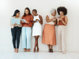 Group Of Five Diverse Young Businesswomen Standing Against A Wall In An Office And Using Tech. Happy Colleagues Talking And Using Technology While Standing In A Row Together At Work