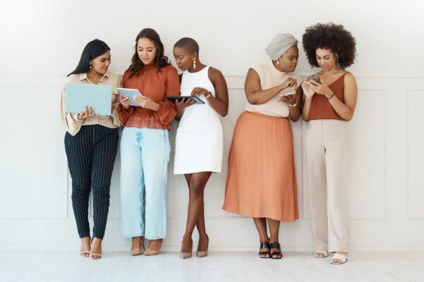 Group Of Five Diverse Young Businesswomen Standing Against A Wall In An Office And Using Tech. Happy Colleagues Talking And Using Technology While Standing In A Row Together At Work