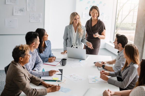 Korean Mature Woman And Caucasian Young Woman Lead Meeting Of Multiracial Work Colleagues Discussing Ideas And Business Together Sitting At Table In Modern Creative Office Interior High Angle View Daytime