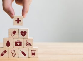A hand arranges building blocks that have images relating to healthcare, including a medical cross, a capsule, a heart, a medical bag, a wheelchair, a stethoscope, a syringe and a pill bottle.