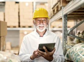 Smiling Senior Worker In Uniform And With Yellow Helmet On Head Holding Tablet And Looking At Camera While Standing In Storage.