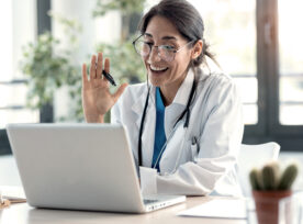Female Doctor Waving And Talking With Colleagues Through A Video Call With A Laptop In The Consultation.