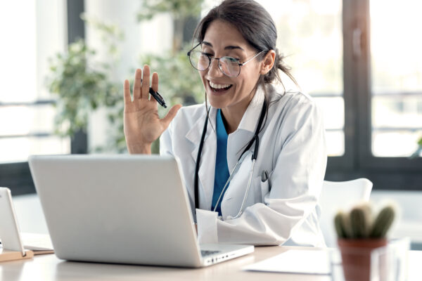 Female Doctor Waving And Talking With Colleagues Through A Video Call With A Laptop In The Consultation.