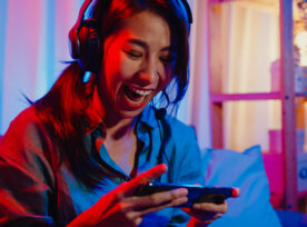 Happy Asia Girl Gamer Wear Headphone Competition Video Game Online With Smartphone Excited Talk With Friend Sit On Couch In Colorful Neon Lights Living Room At Home.