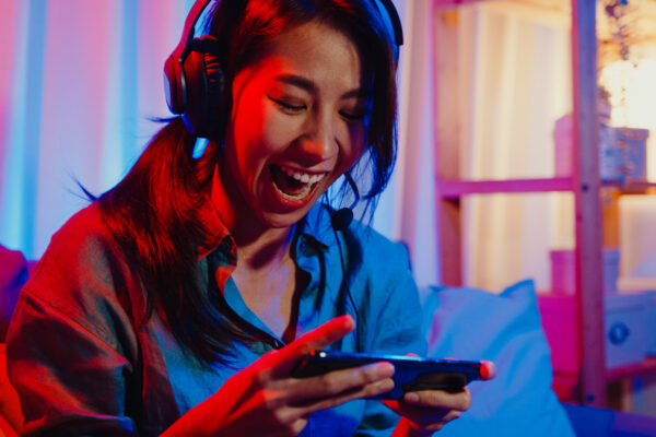 Happy Asia Girl Gamer Wear Headphone Competition Video Game Online With Smartphone Excited Talk With Friend Sit On Couch In Colorful Neon Lights Living Room At Home.