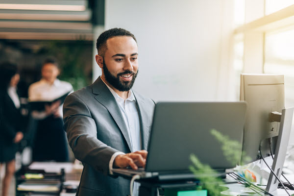 Multiracial Portuguese Jamaican mid adult businessman with beard standing smiling at desk with laptop reviewing data in bright business office wearing suit