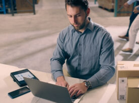 Laptop, Industry And Man In Logistics Warehouse Doing Research For Stock Inventory With Boxes. Technology, Computer And Male Industrial Worker Typing And Working On Ecommerce Report In Cargo Factory.