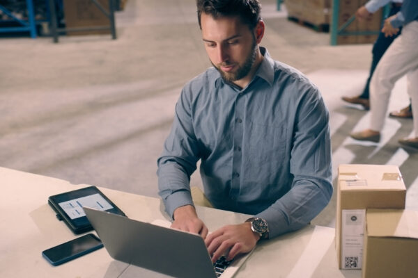 Laptop, Industry And Man In Logistics Warehouse Doing Research For Stock Inventory With Boxes. Technology, Computer And Male Industrial Worker Typing And Working On Ecommerce Report In Cargo Factory.