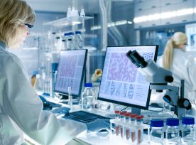 Senior Female Scientist Works With High Tech Equipment In A Modern Laboratory. Her Colleagues Are Working Beside Her.