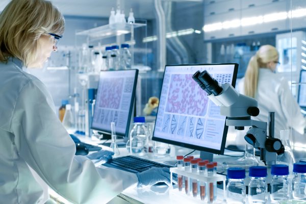 Senior Female Scientist Works With High Tech Equipment In A Modern Laboratory. Her Colleagues Are Working Beside Her.