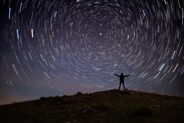 Long exposure night photo of the silhouette of a person on a hill with the stars in the sky tracking as radial lines around the North Star. Symbolizing goal setting.