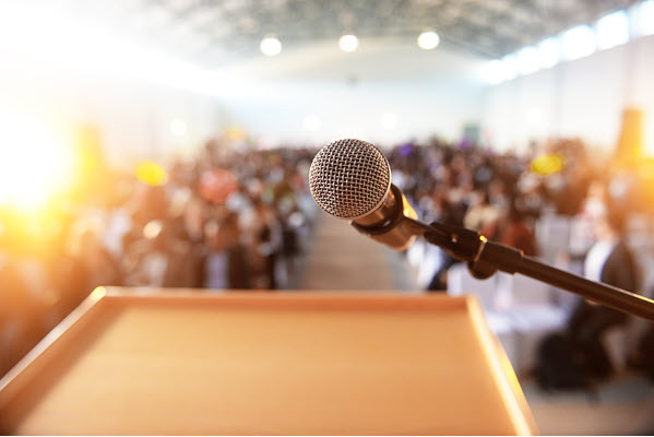 Speaker’s lectern with microphone in front of a blurry audience.