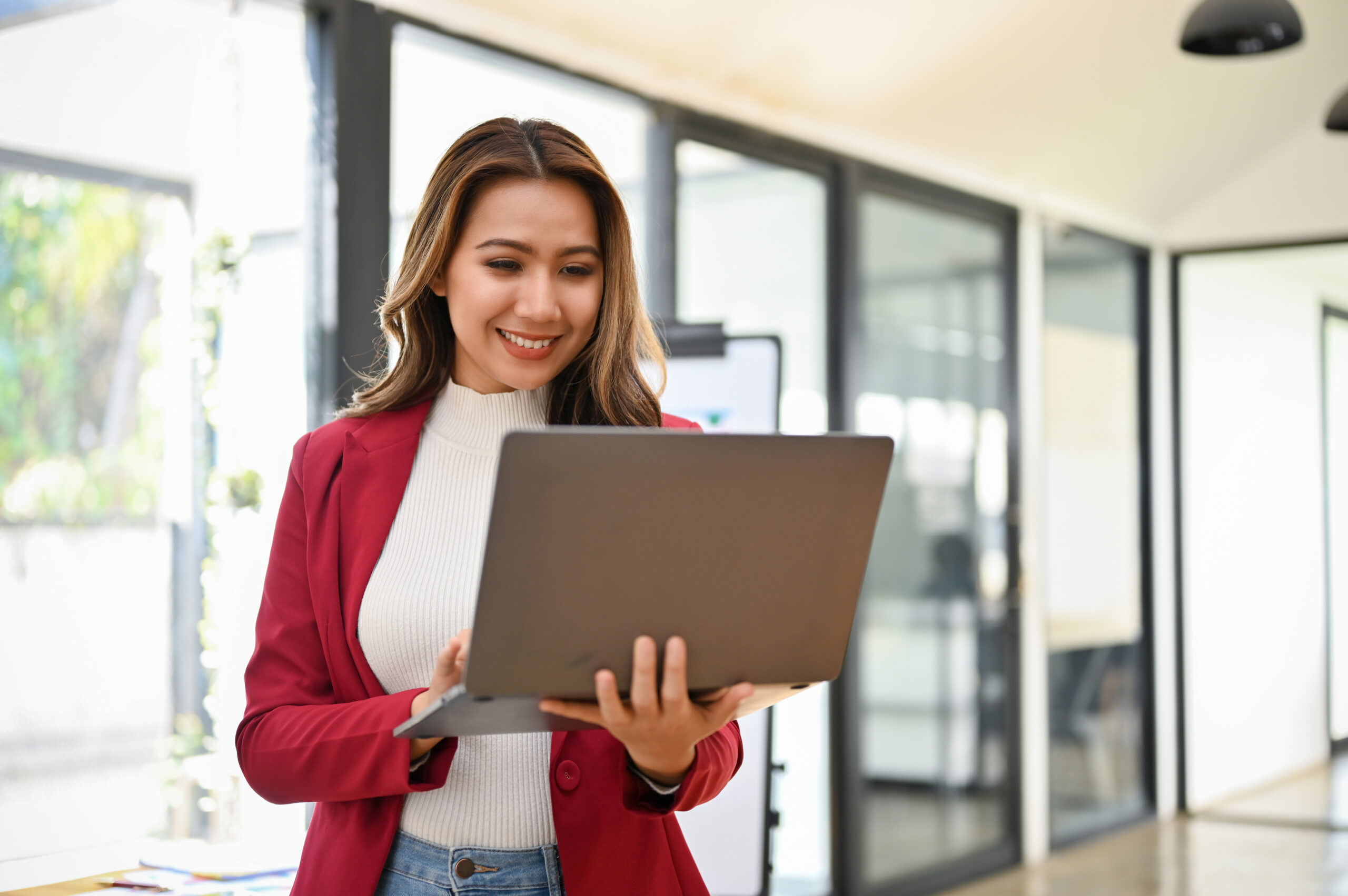 Professional Millennial Asian Businesswoman Stands In Her Office Using Portable Laptop